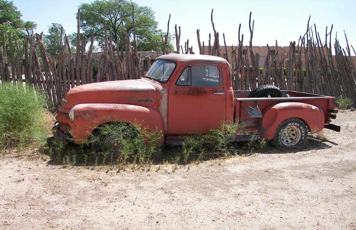 Rusty red vintage truck partially covered in weeds, parked beside a wooden fence